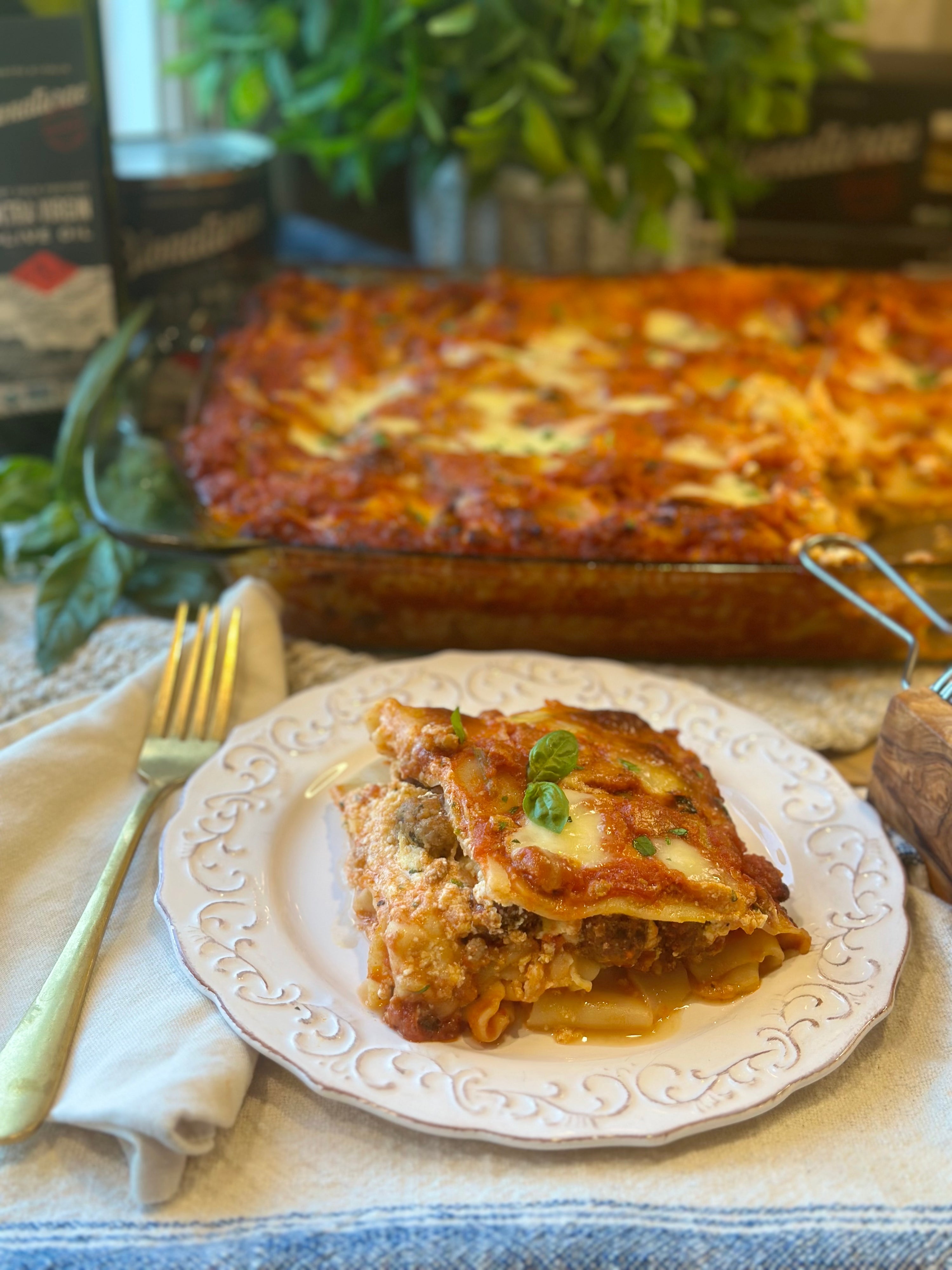 Slice of Neapolitan-style lasagna on a white decorative plate with a glass baking dish of lasagna and basil in the background