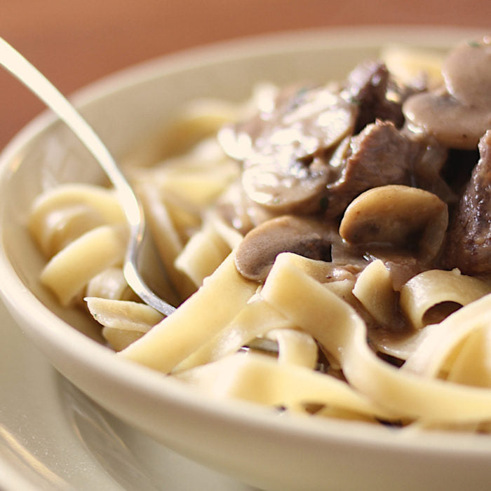 Close up of bowl of gluten free beef stroganoff showing brown rice tagliatelle, sliced mushrooms and beef in creamy sauce