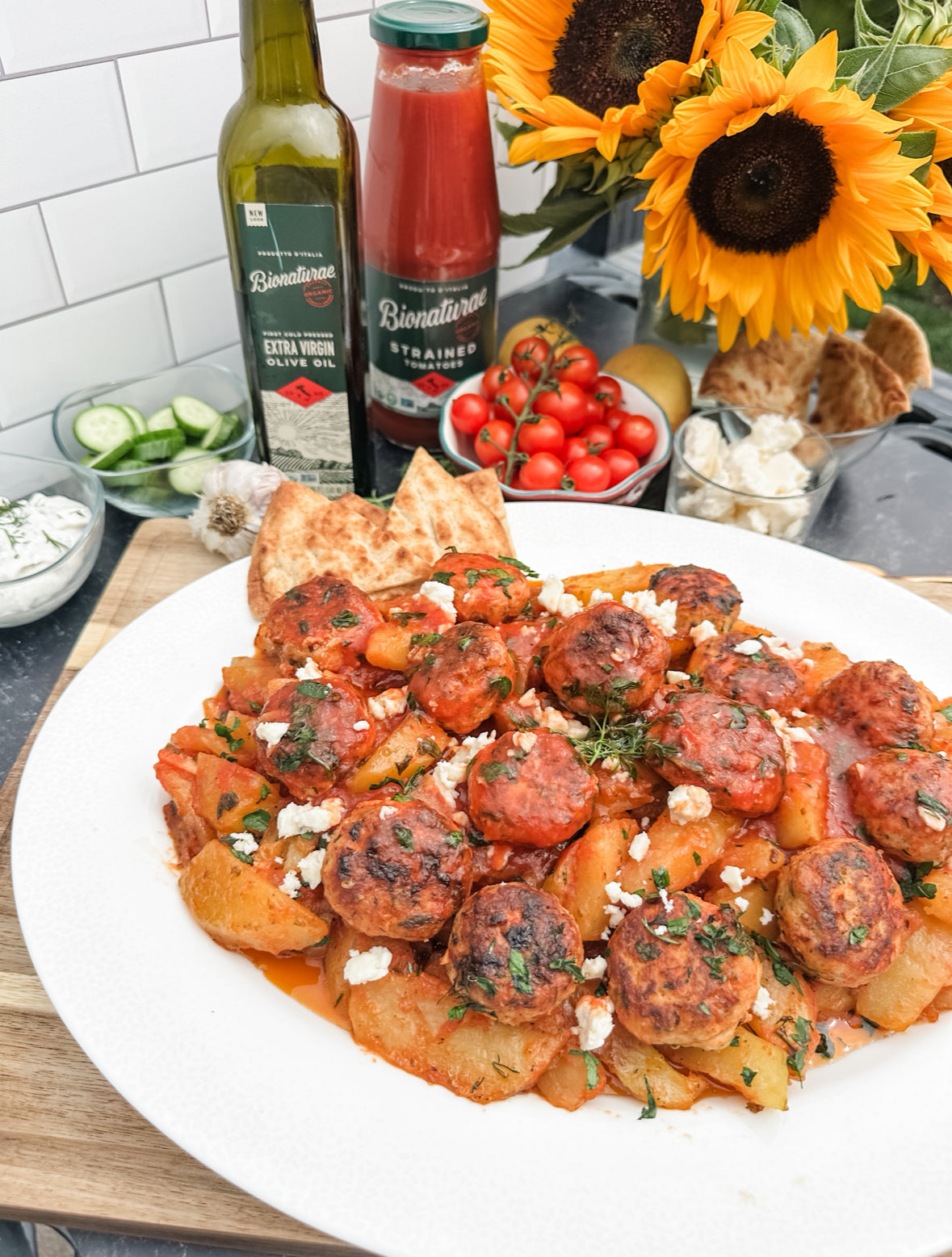 Greek chicken meatballs and potatoes plated with crumbled feta, pita, and bottles of olive oil and strained tomatoes in the background