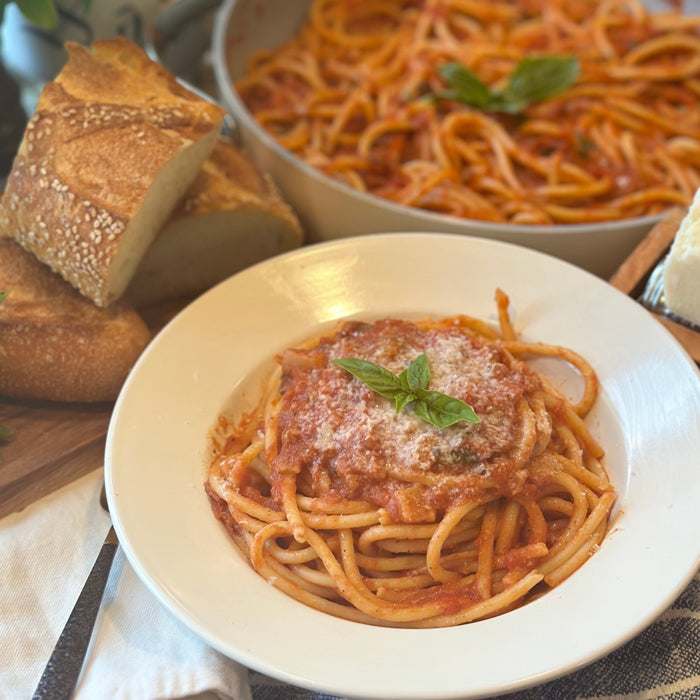 Bucatini Amatriciana in a white bowl topped with tomato sauce, grated cheese and basil, with bread and a pan of pasta behind