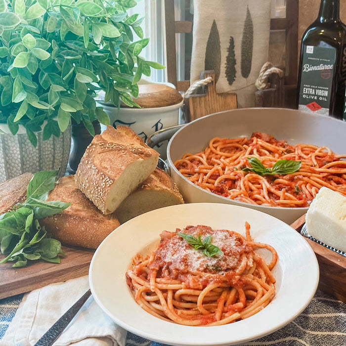 Bucatini amatriciana in a white bowl topped with basil and grated cheese, on a table with crusty bread and a bottle of olive oil