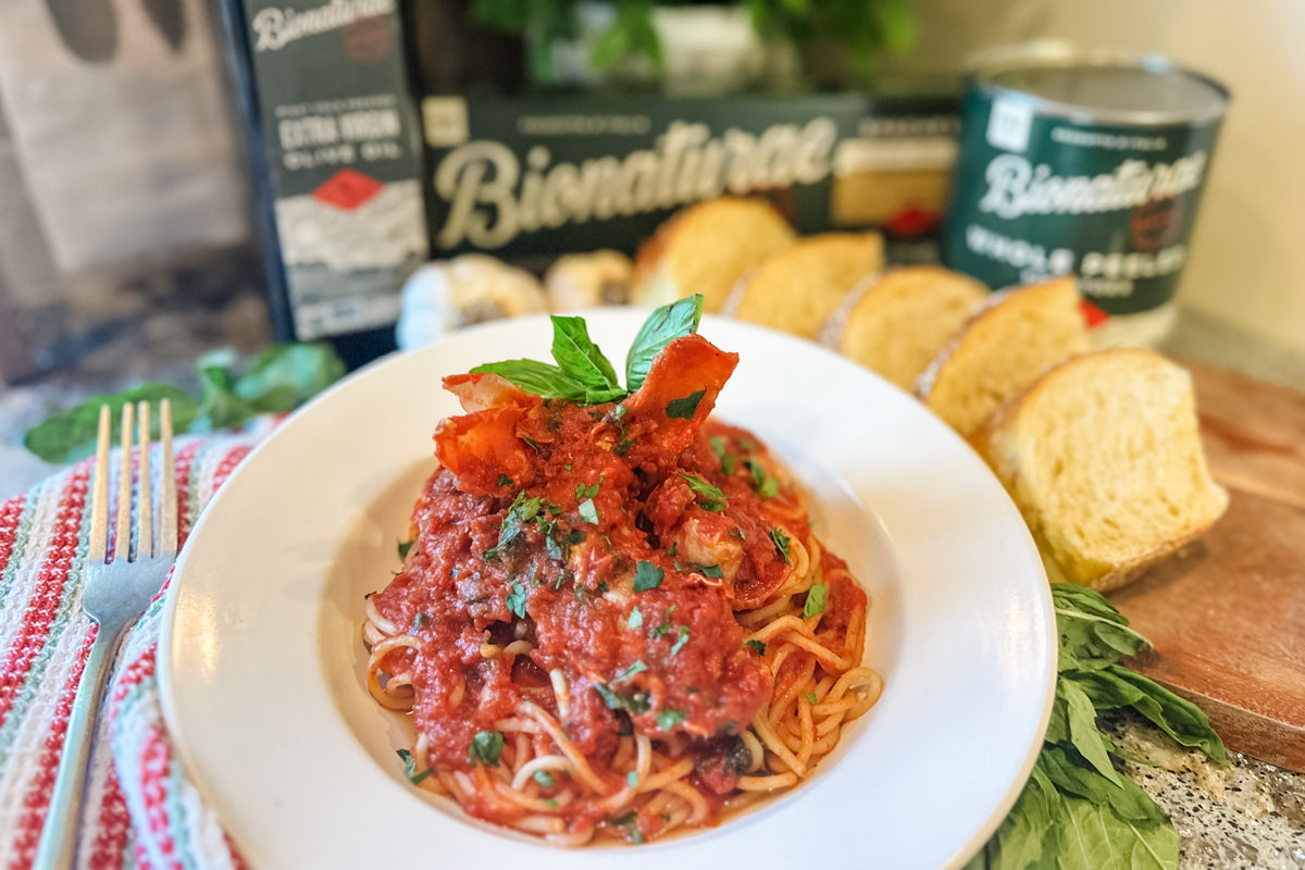 Spaghetti in lobster tomato sauce topped with basil, served in a white bowl with sliced bread and Bionaturae cans in the background
