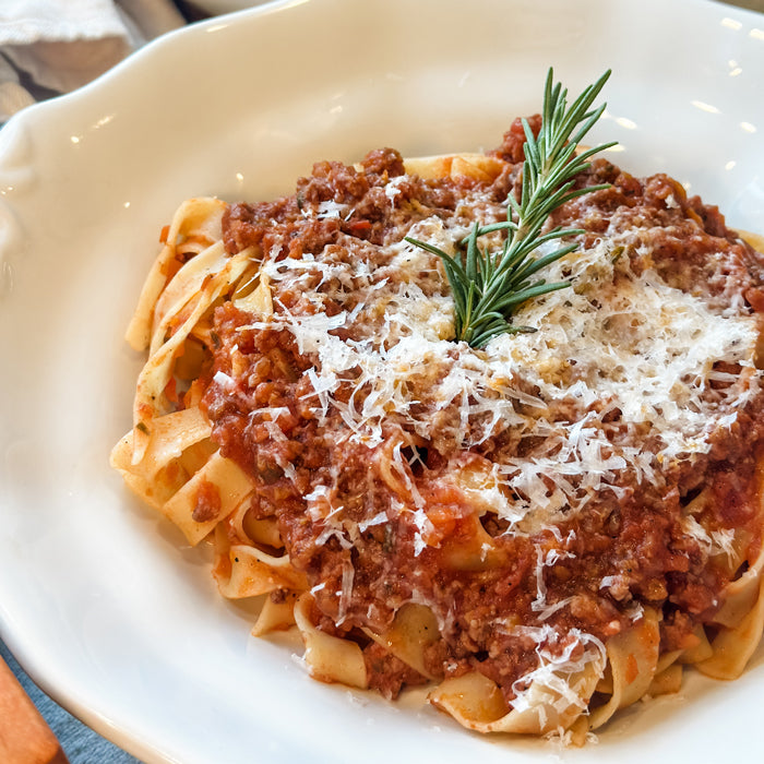 Bowl of tagliatelle topped with Tuscan ragu, grated Parmesan, and a rosemary sprig on a white plate
