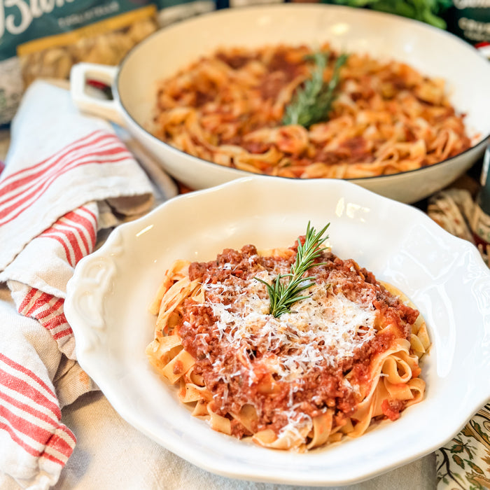 Two bowls of pasta with Tuscan ragu topped with grated cheese and a rosemary sprig on a linen cloth with a serving pan behind