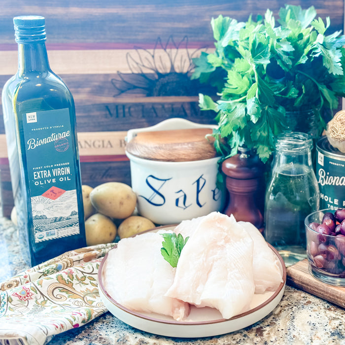 Plate of raw white fish fillets with parsley, potatoes, olives, salt jar, and Bionaturae extra virgin olive oil