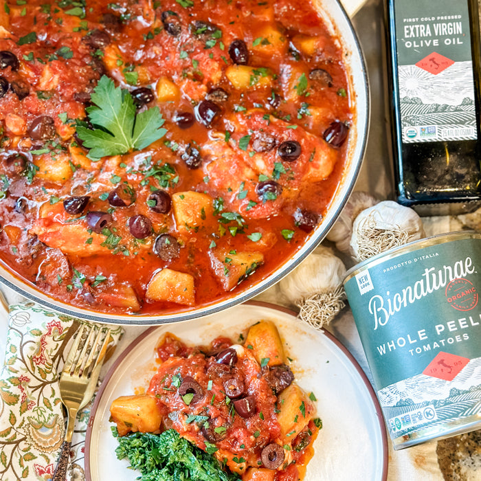 Skillet of Acqua Pazza poached fish in tomato sauce with olives, beside Bionaturae tomatoes and olive oil