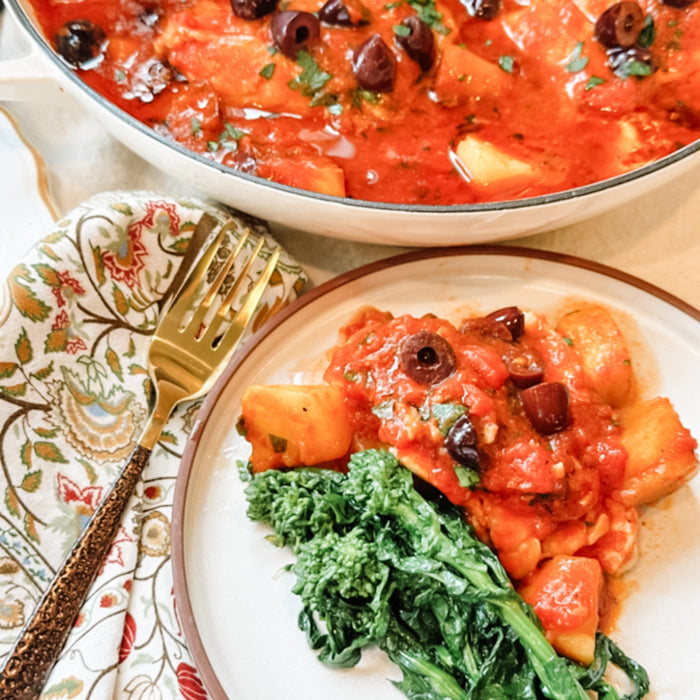 Acqua Pazza (Italian poached fish) with tomato-potato sauce, olives, and sauteed greens on a plate beside a skillet