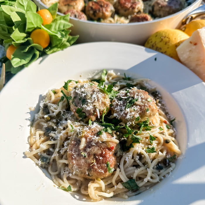 Chicken piccata meatballs served over capellini with grated Parmesan and parsley on a white plate, pan and lemons in sunlight