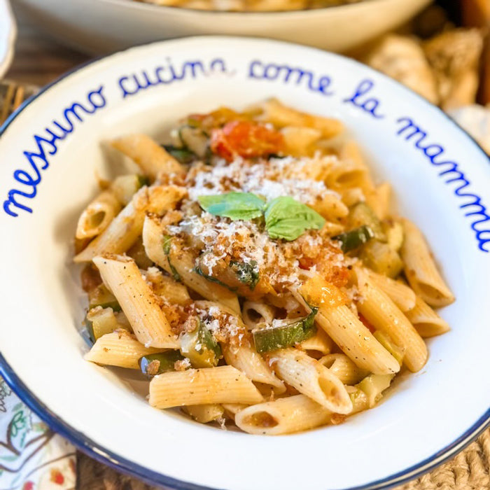 Overhead view of penne with zucchini, tomatoes, cheese and basil in a white bowl with rim text nessuno cucina come la mamma