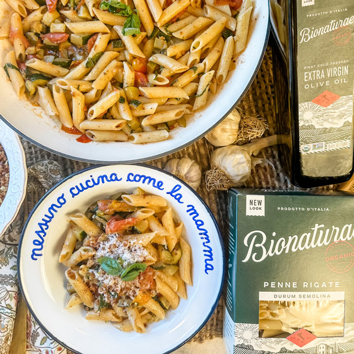 Bowl of penne with zucchini, tomatoes, basil and grated cheese next to Bionaturae penne box and olive oil bottle