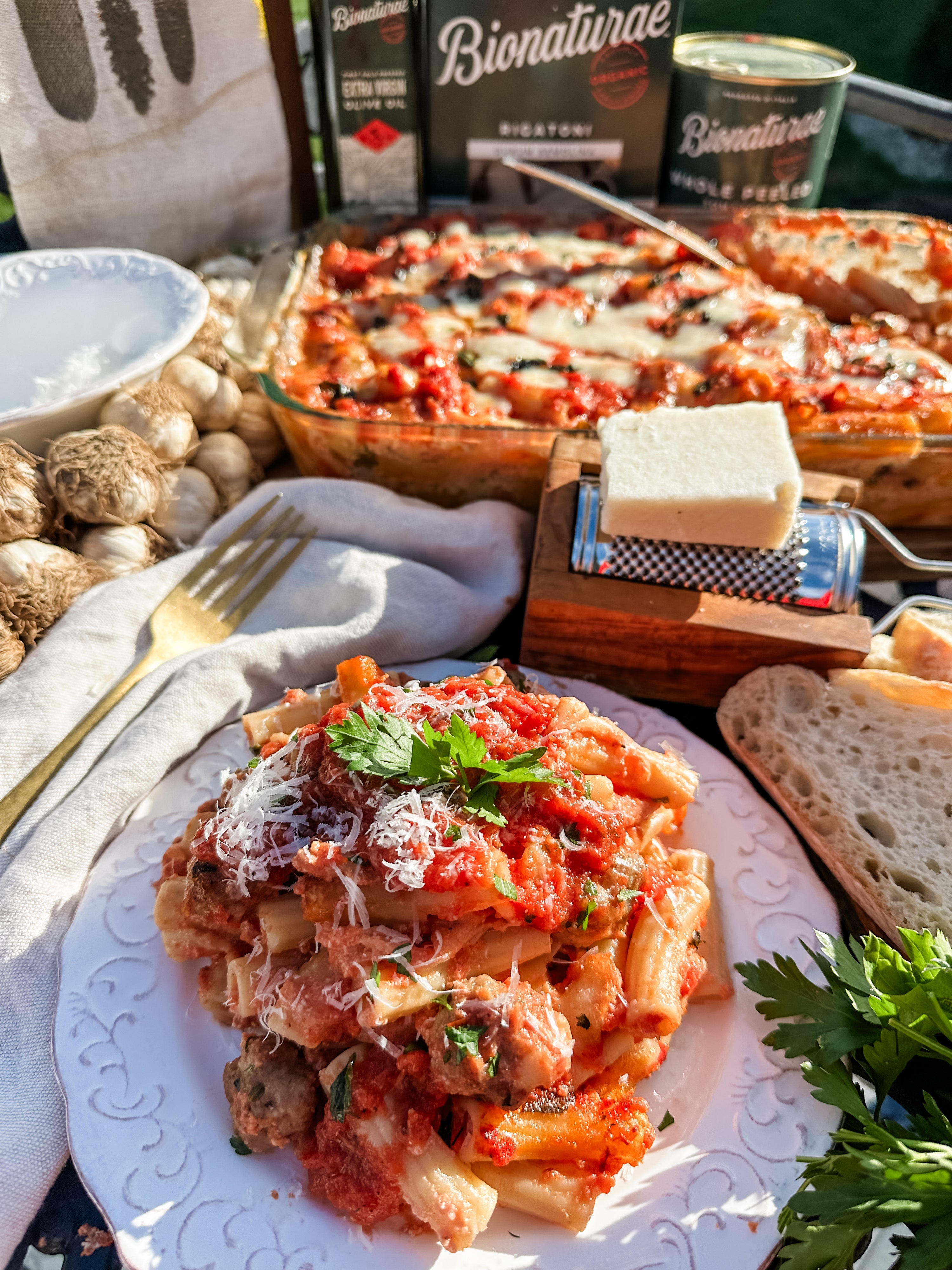 Baked rigatoni with mini sausage meatballs on a plate with grated cheese, bread, and the baking dish in a rustic setting