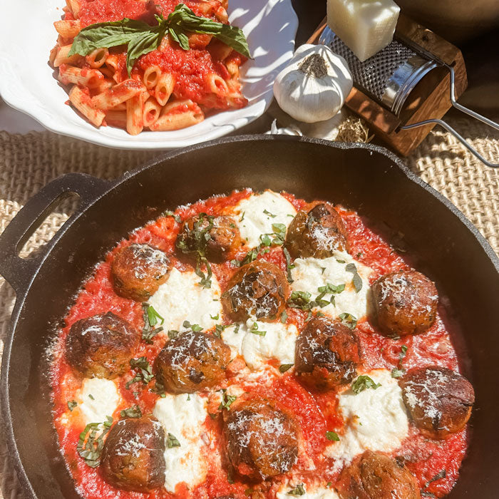 Eggplant meatballs simmering in homemade marinara with melted ricotta in a cast-iron skillet, with a plate of pasta and garlic