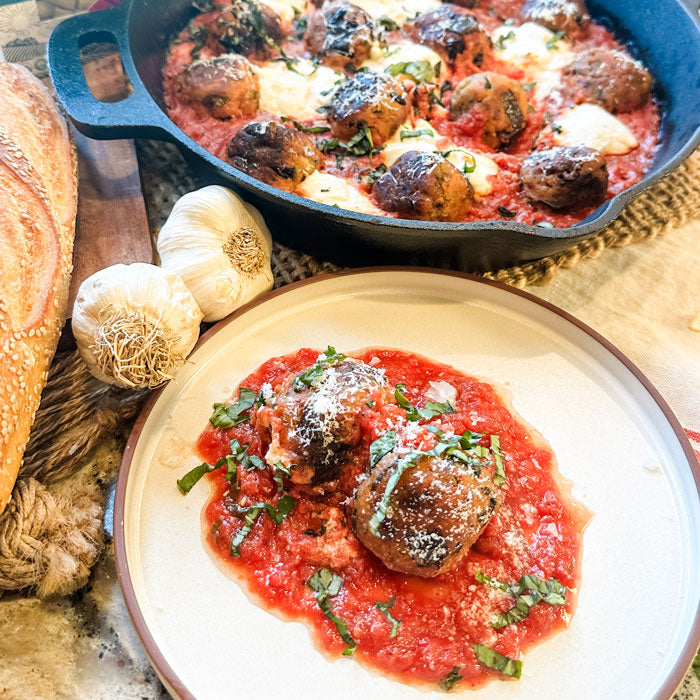 Eggplant meatballs in homemade marinara on a white plate, with a cast-iron skillet of more meatballs, garlic and crusty bread behind