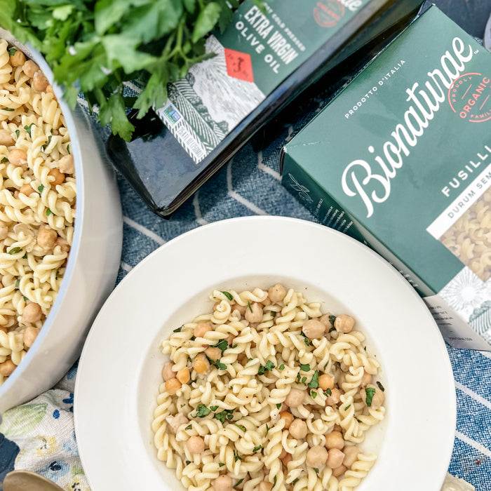 Bowl of Italian fusilli pasta and chickpeas served with parsley, beside an olive oil bottle and pasta box on a blue cloth