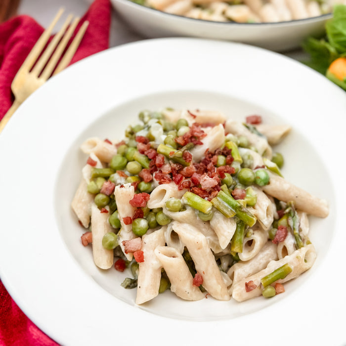 Overhead view of Sourdough Penne with asparagus, peas, and crispy pancetta in a creamy sauce on a white plate with a red napkin