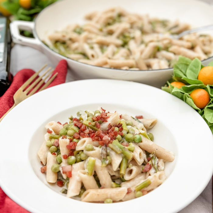Sourdough penne with asparagus, peas, and crispy pancetta in a white bowl with a serving pan and citrus on the table