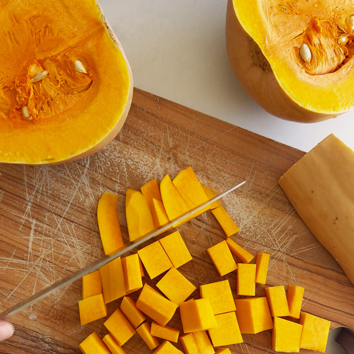 Butternut squash being diced with a chef’s knife on a wooden cutting board, halved squash pieces visible for autumn soup