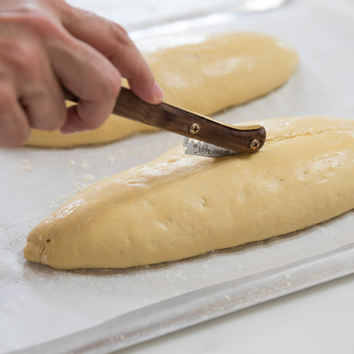 Two loaves of Einkorn Italian bread dough being scored with a razor on a parchment-lined baking sheet