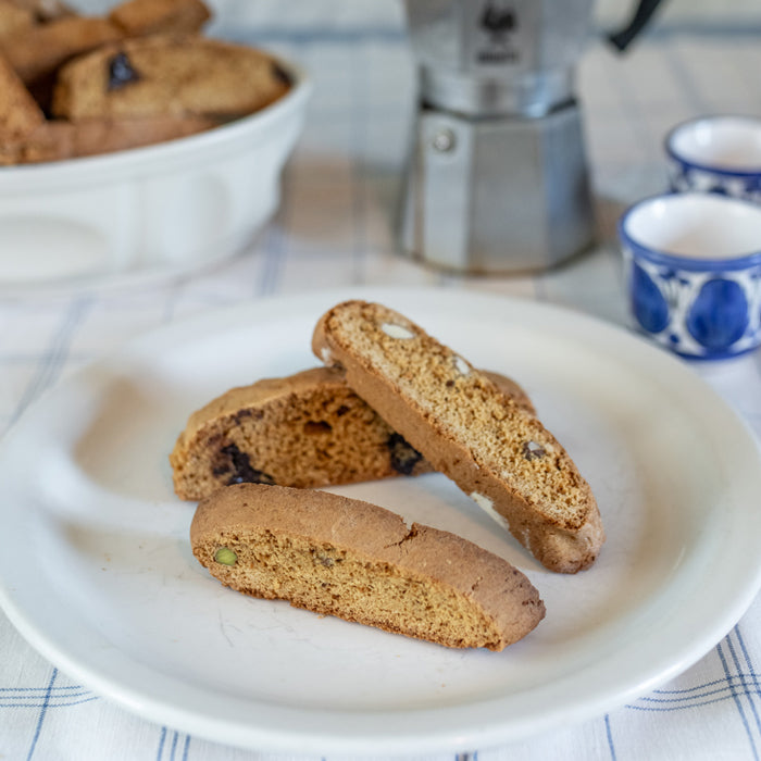 Three sliced Einkorn biscotti showing pistachios and almonds on a white plate with a moka pot and blue espresso cups behind