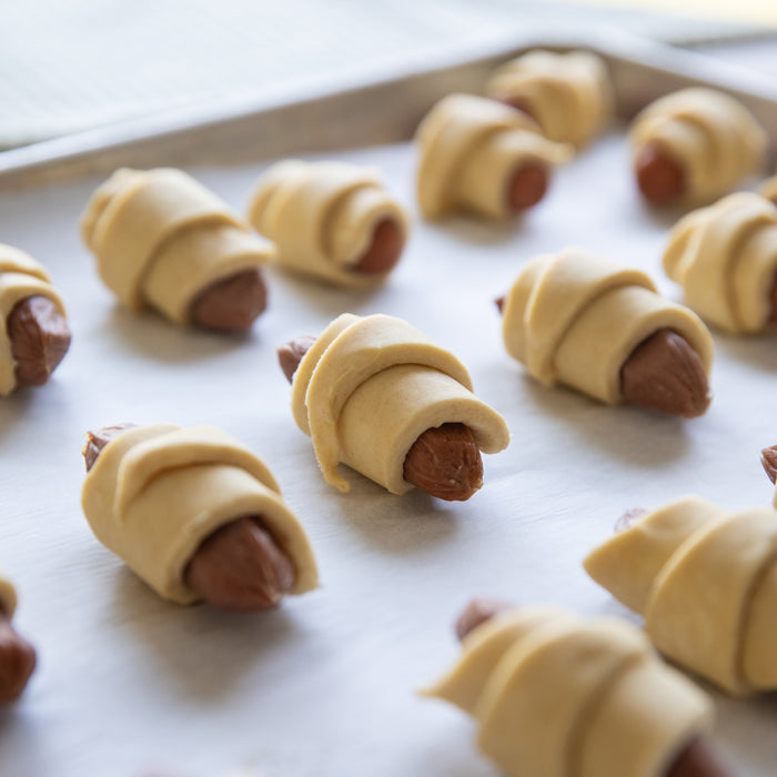 Close-up of unbaked mini pigs in a blanket made with einkorn pastry wrapped around mini beef franks on a baking sheet