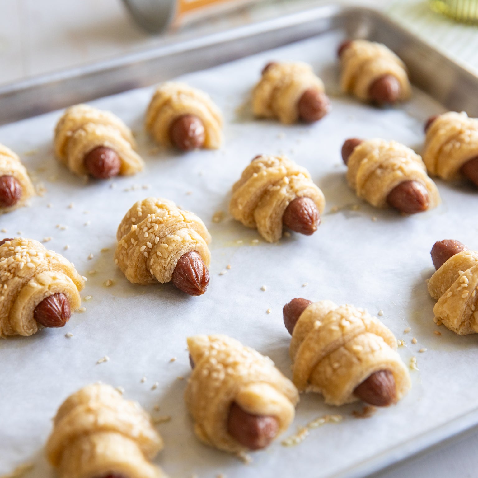 Mini pigs in a blanket made with einkorn puff pastry on a parchment-lined baking sheet, sprinkled with sesame seeds