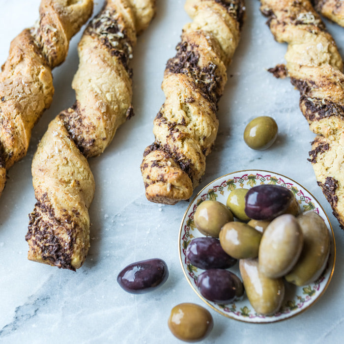 Top view of Einkorn grissini twisted breadsticks swirled with olive tapenade beside a small dish of mixed olives on marble
