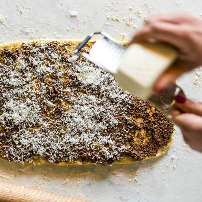 Rolled einkorn dough spread with olive tapenade while someone grates cheese over it on a floured surface