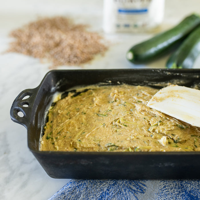 Zucchini bread batter in a buttered loaf pan with a spatula, einkorn flour and zucchinis blurred in the background