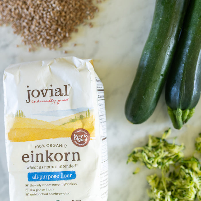 Angled view of Jovial 100 percent organic einkorn all-purpose flour bag beside whole and shredded zucchini on a marble countertop