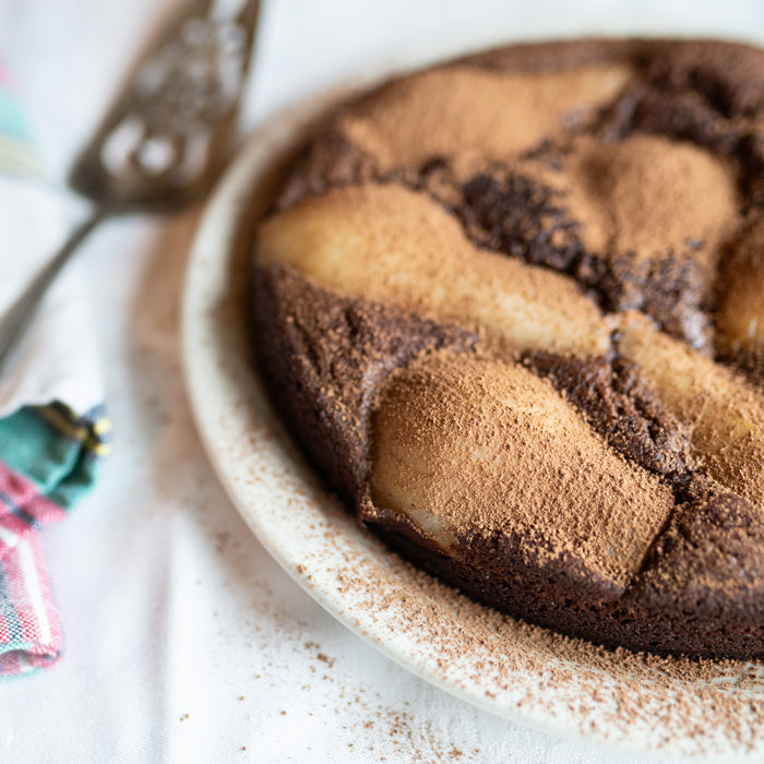 Angled close-up of chocolate pear pudding cake showcasing pear halves and cocoa-dusted top in a white pie dish on white cloth