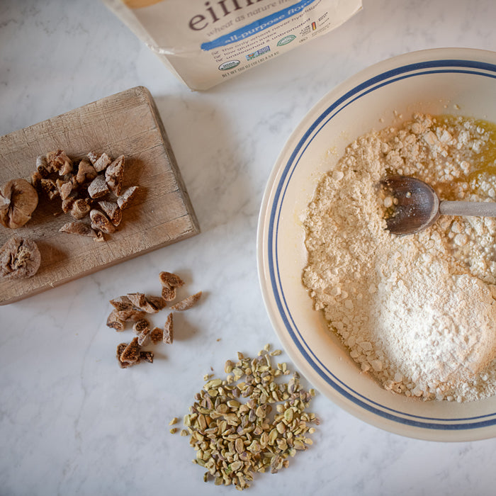 Bowl of einkorn flour mixed with egg and oil next to chopped figs and shelled pistachios on a marble counter