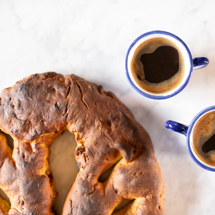 Top-down of Einkorn breakfast bread with figs and pistachios showing golden crust beside two espresso cups on marble