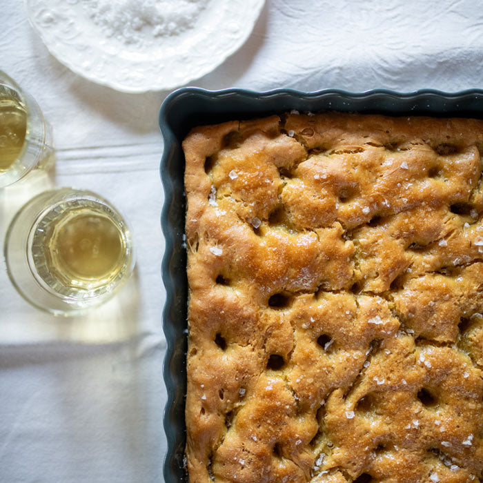 Top-down view of rectangular einkorn focaccia in a scalloped pan, dimpled golden crust sprinkled with sea salt on a tablecloth