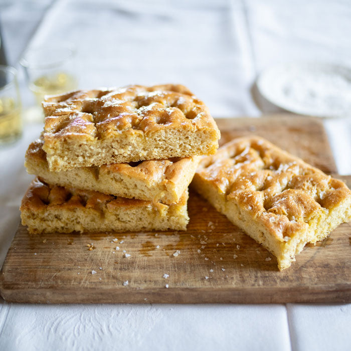 Stacked slices of Einkorn focaccia on a wooden board showing dimpled golden crust, coarse sea salt, and olive oil sheen