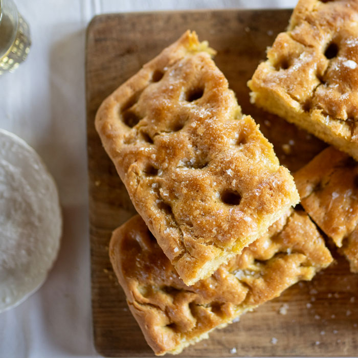 Overhead view of Einkorn focaccia cut into squares showcasing a golden dimpled crust sprinkled with coarse sea salt on a wooden board