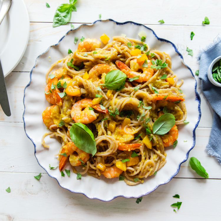 Organic cassava spaghetti tossed with sautéed shrimp, diced yellow peppers, and fresh basil on a white scalloped plate, overhead view