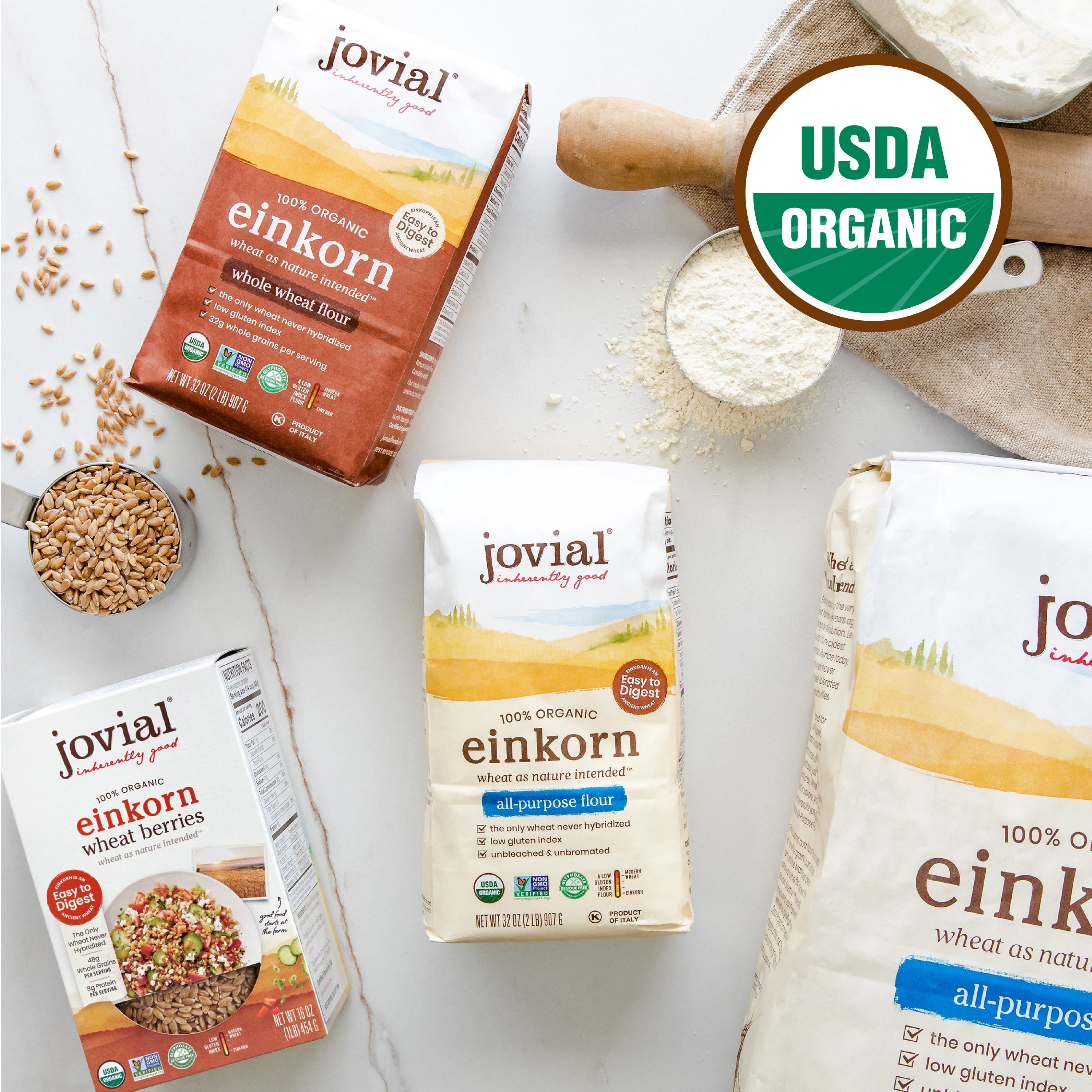 Top-down of Jovial Einkorn flour and wheat berries packages with USDA Organic badge and scoop on marble surface