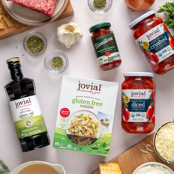 Top-down view of a Jovial gluten free brown rice mafalda pasta box surrounded by olive oil, jars of crushed and diced tomatoes, garlic, herbs, and grated cheese on a light countertop