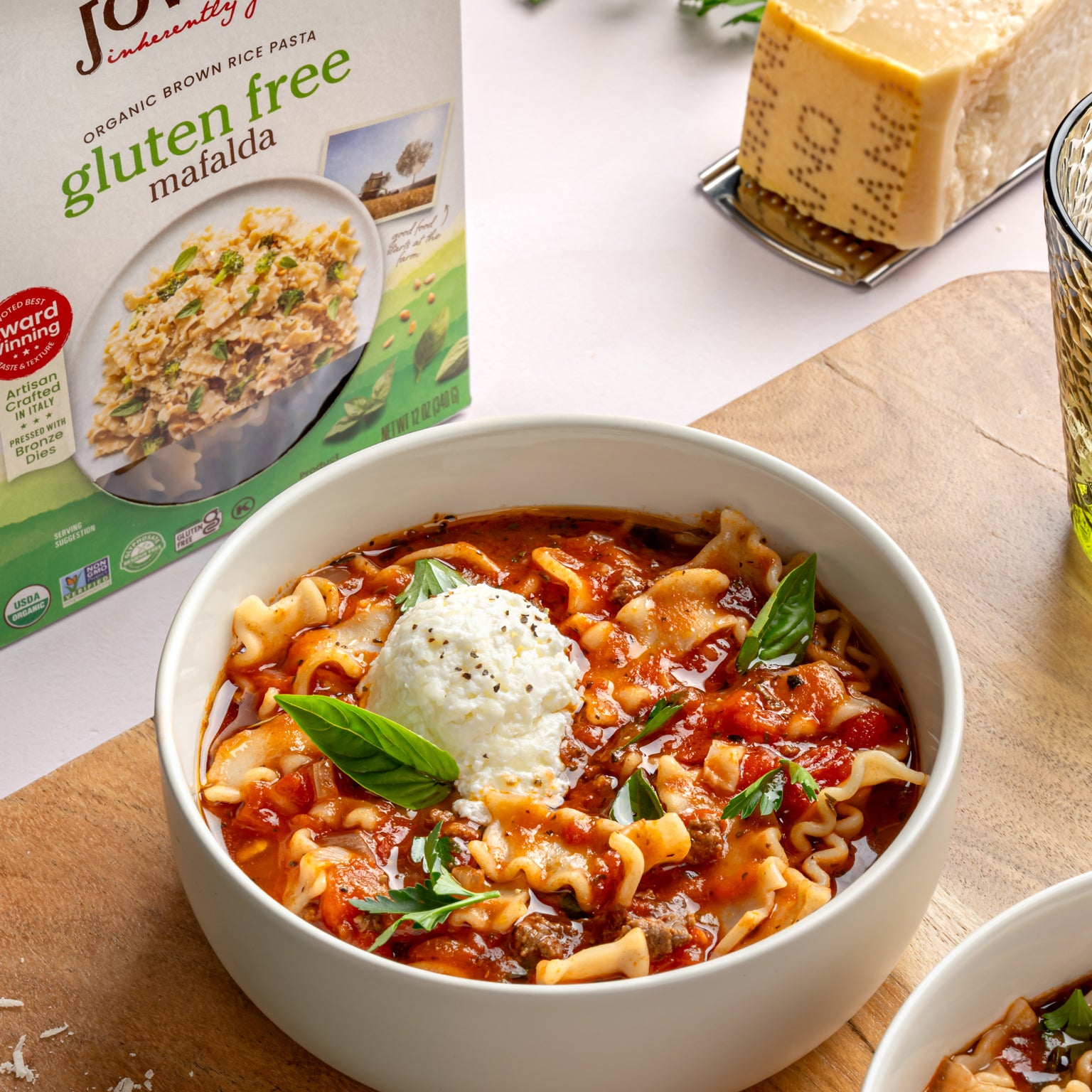 Close-up of lasagna soup with gluten-free brown rice mafalda showcasing a ricotta dollop and basil garnish with product box and cheese in the background