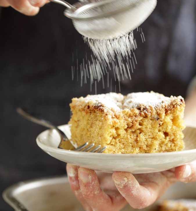 Slice of overnight Einkorn kefir crumb cake being dusted with powdered sugar on a small plate held by flour-dusted hands
