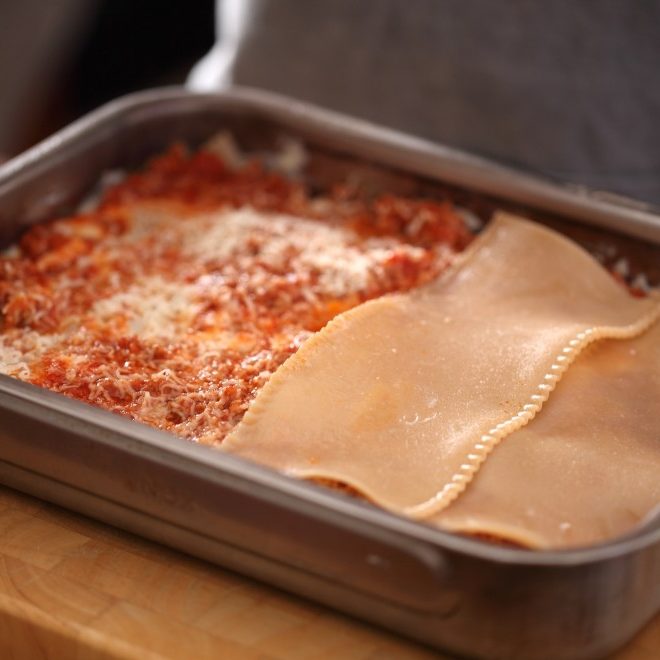 Gluten-free lasagna being layered in a metal baking pan with Bolognese sauce and uncooked pasta sheets