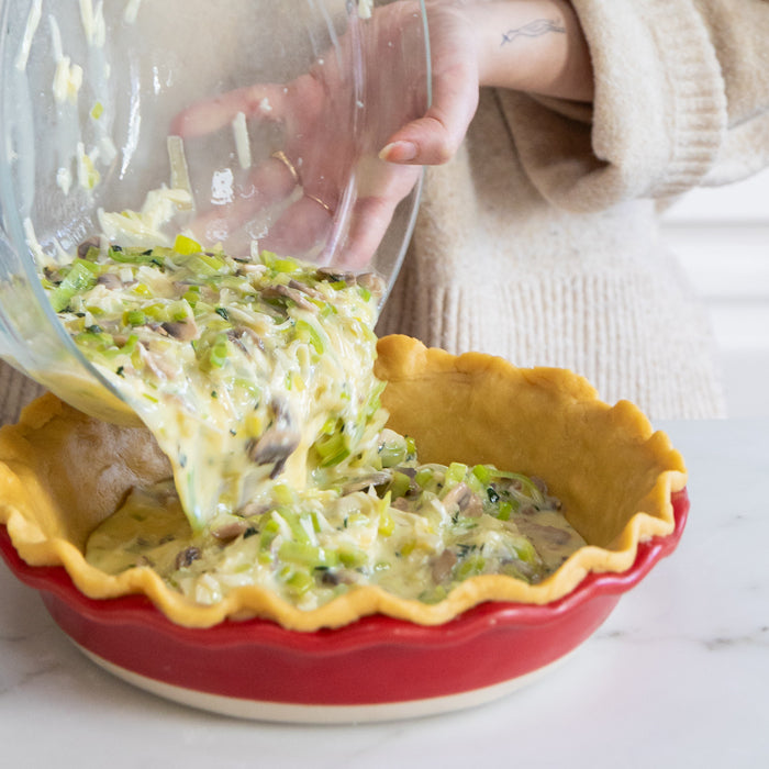 Hands pouring leek and mushroom egg filling into an einkorn pastry crust in a red pie dish on a marble counter