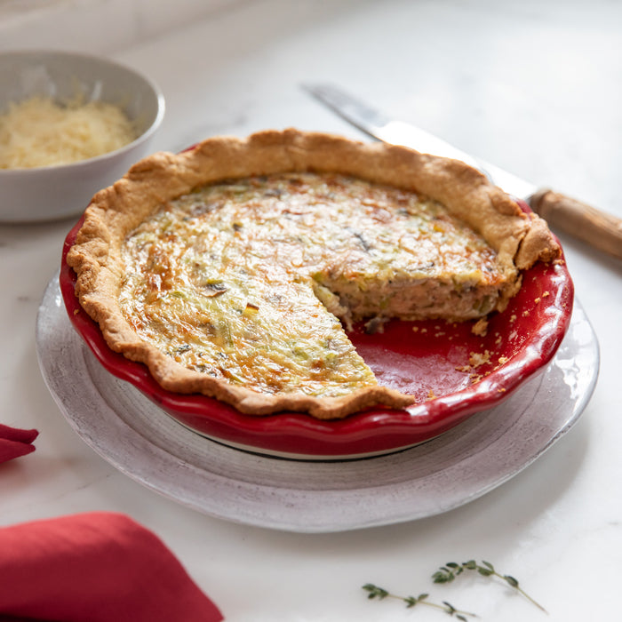 Three-quarter view of leek and mushroom Einkorn quiche in a red pie dish with one slice removed on a marble counter