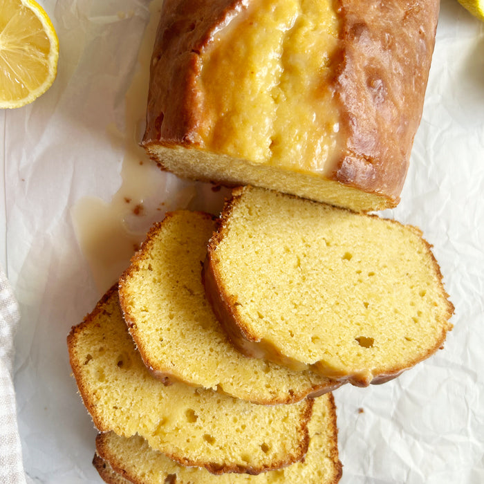 Top-down view of sliced Einkorn sourdough lemon yogurt loaf showcasing moist yellow crumb and lemon glaze on parchment with halved lemon