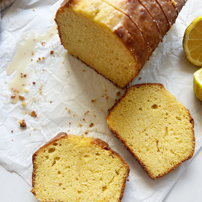 Top-down view of sliced einkorn lemon yogurt loaf showcasing moist crumb and lemon glaze on crumpled parchment with lemon halves