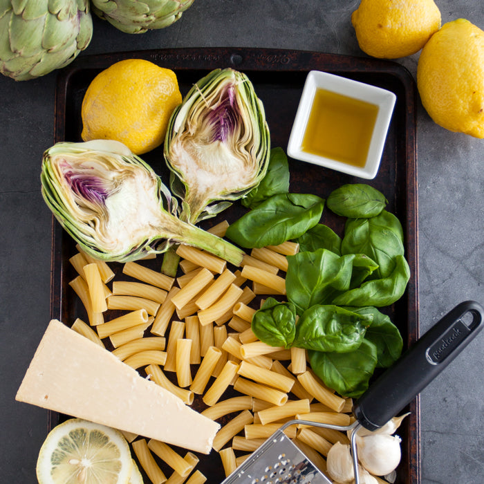 Top-down view of rigatoni pasta with halved artichokes, basil, lemon, Parmesan wedge, garlic and olive oil on a dark tray