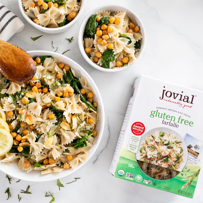 Bowl of lemon spinach farfalle with crispy chickpeas and lemon slice beside a Jovial gluten free brown rice farfalle box on a marble countertop