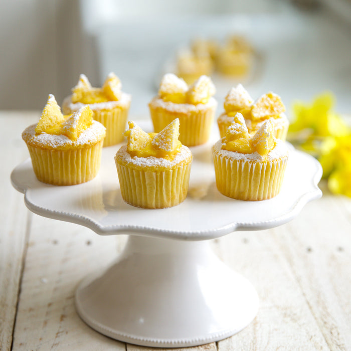 Close-up of six Einkorn lemon cupcakes on a white cake stand topped with lemon curd and powdered sugar, soft blurred kitchen background