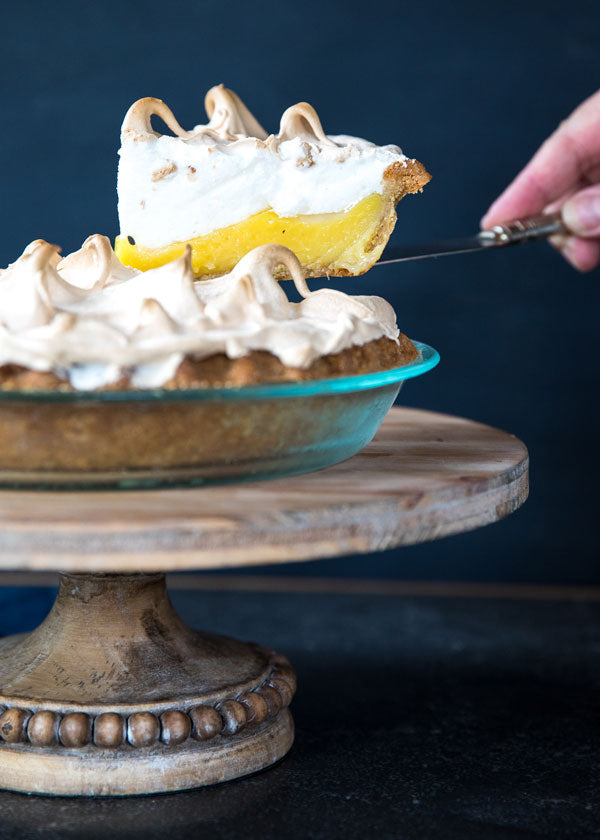 Side view of lemon meringue pie slice showing lemon curd and toasted meringue on glass pie plate on wooden cake stand