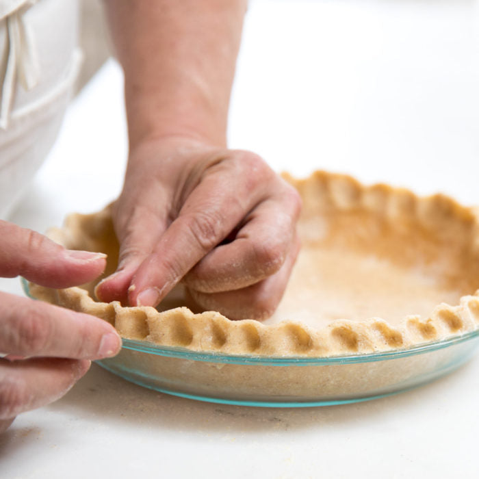 Hands crimping an unbaked Einkorn pie crust in a glass pie dish on a white countertop